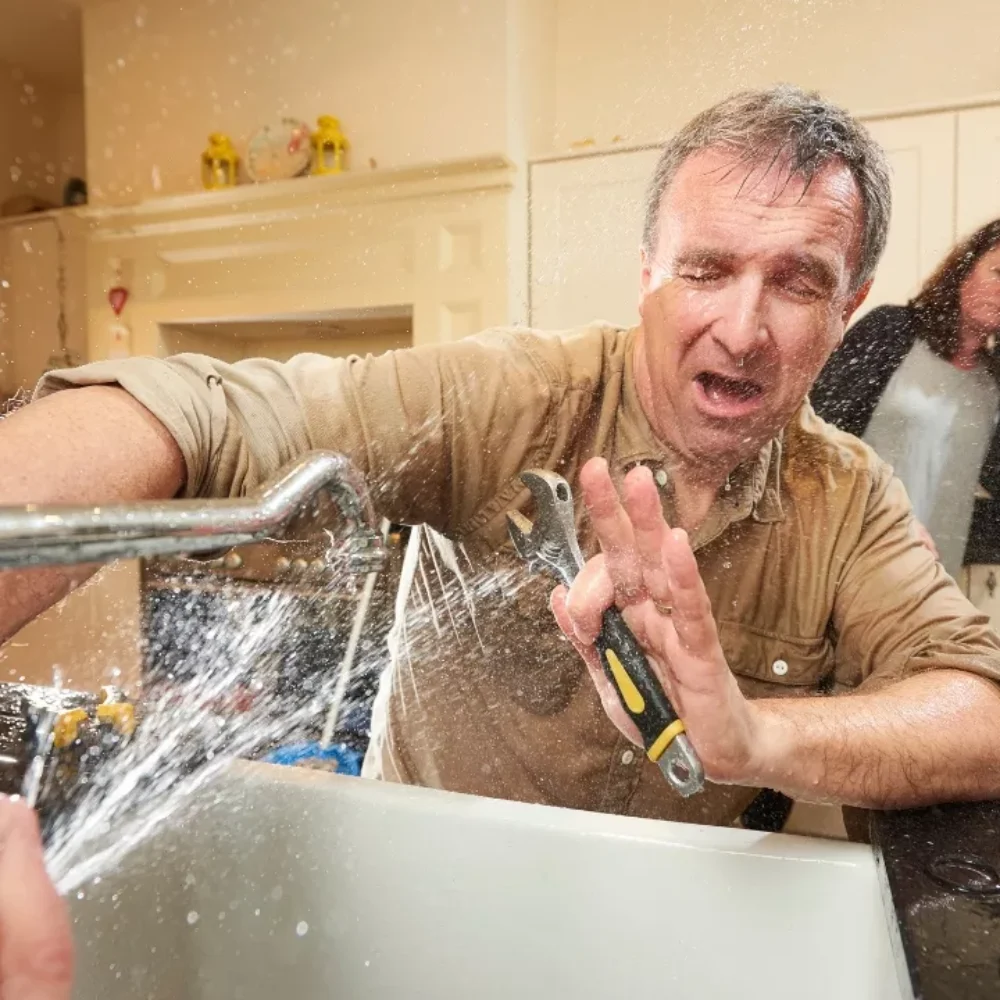 man trying to fix a sink with water spraying in his face while a woman is on the phone calling for help