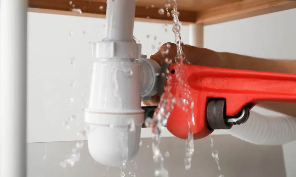 Person under sink with a red pipe wrench performing plumbing maintenance to stop water spraying